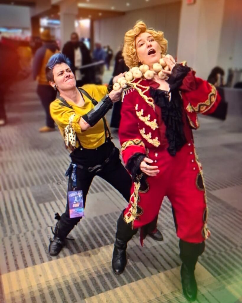 Char Grey dressed as Stede Bonnet in the cursed suit being choked by garlic by a Jim cosplayer